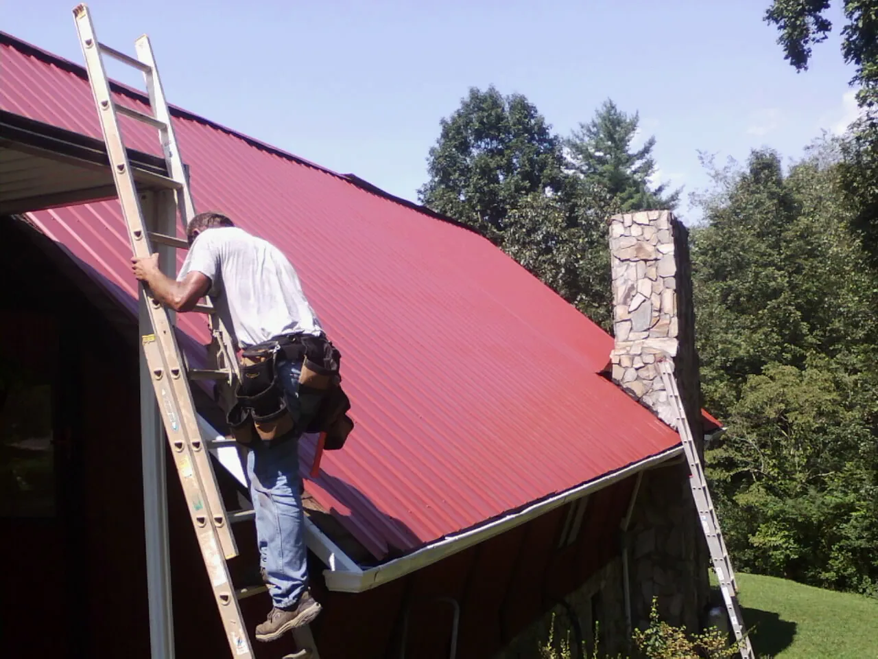 Roofer on a ladder beside a red metal roof and stone chimney — Soffit & Fascia Repair by Roofing Brothers in East Canton